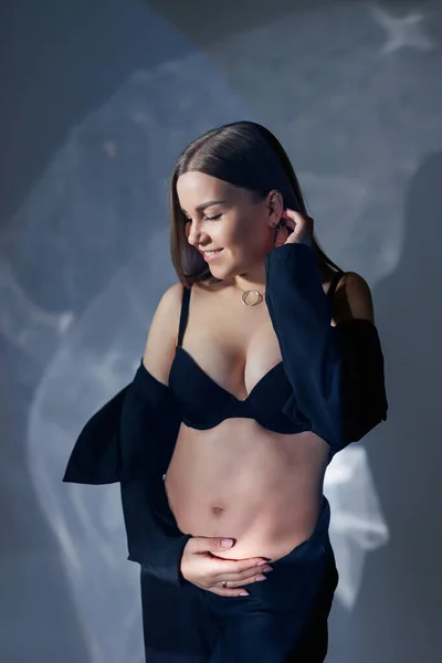 黒の古典的なスーツの若い妊婦の肖像画 灰色の背景に妊娠中の女性は彼女の腹を抱擁 — ストック写真