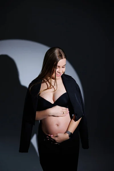 stock image Happy pregnant woman in a black suit on a gray background. Beautiful elegant pregnant woman. Gentle studio photos of a pregnant woman