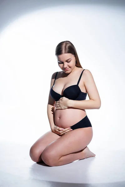 黒の下着姿の可愛い妊婦さんが白い背景に腰を下ろしています 妊婦用の女性用下着 — ストック写真