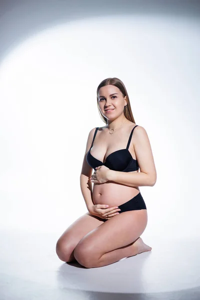 黒の下着姿の可愛い妊婦さんが白い背景に腰を下ろしています 妊婦用の女性用下着 — ストック写真