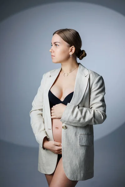 白い背景のスタジオには 長いジャケットと下着を着たスリムな妊婦が立っており 彼女の胃に手をつないでいます 妊婦用の女性用下着 — ストック写真