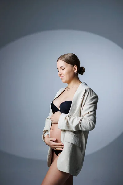 白い背景のスタジオには 長いジャケットと下着を着たスリムな妊婦が立っており 彼女の胃に手をつないでいます 妊婦用の女性用下着 — ストック写真