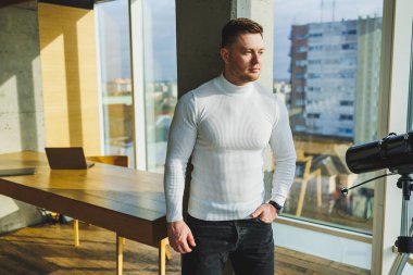 Beyaz kazaklı genç yakışıklı bir adam ferah, parlak bir ofiste duruyor. Modern başarılı insan kavramı. Açık ofis alanında gülümseyen genç adam.