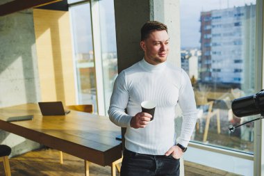 Parlak çalışma alanında uzaktan çalışırken kahve içen genç ve tatlı bir serbest çalışan. Beyaz kazaklı ve pantolonlu bir adam ofiste mutlu mesut çalışır.