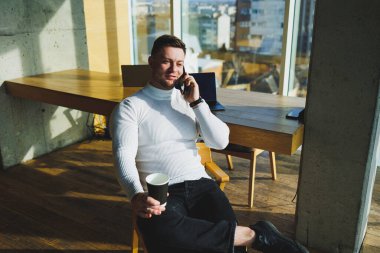 Parlak çalışma alanında uzaktan çalışırken kahve içen genç ve tatlı bir serbest çalışan. Beyaz kazaklı ve pantolonlu bir adam ofiste mutlu mesut çalışır.