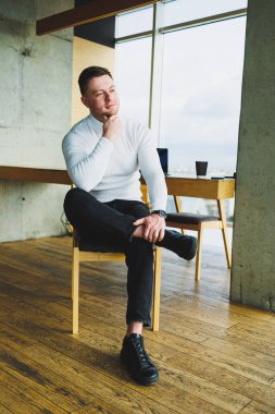 Beyaz kazak ve pantolon giyen çekici genç bir adam parlak bir ofiste otururken mutlu bir şekilde gülümsüyor. Rahat giyimli bir adam.