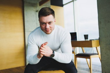 Beyaz kazak ve pantolon giyen çekici genç bir adam parlak bir ofiste otururken mutlu bir şekilde gülümsüyor. Rahat giyimli bir adam.