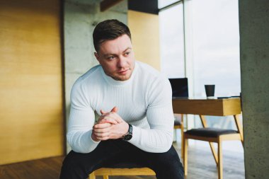 Beyaz kazak ve pantolon giyen çekici genç bir adam parlak bir ofiste otururken mutlu bir şekilde gülümsüyor. Rahat giyimli bir adam.