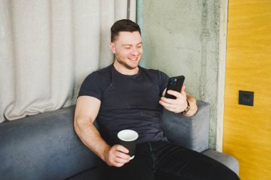 Tişörtlü başarılı genç adam koltukta oturuyor, telefon ediyor, kahve içiyor, serbest olarak çalışıyor, dizüstü bilgisayara bakıyor ve gülümsüyor.