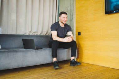 Tişörtlü yakışıklı genç adam kanepede oturuyor ve kahve içiyor, başlangıçta bağımsız olarak çalışıyor, dizüstü bilgisayarına bakıyor ve gülümsüyor.