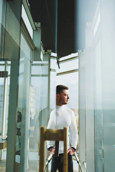 A young man in a modern workspace stands near a large window. A man in casual clothes with a laptop in his hands. Remote work. Nice young manager