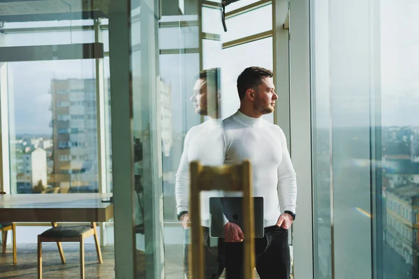 A young man in a modern workspace stands near a large window. A man in casual clothes with a laptop in his hands. Remote work. Nice young manager