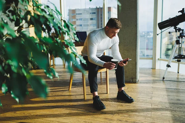 stock image Cute young male freelancer drinking coffee while sitting in bright workspace working remotely. A man in a white sweater and trousers works happily in the office