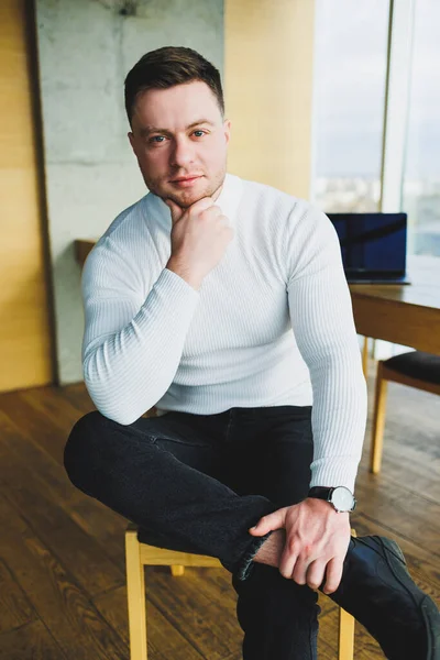 An attractive young man in a white sweater and trousers is smiling contentedly while sitting in a bright office. Man in comfortable casual clothes