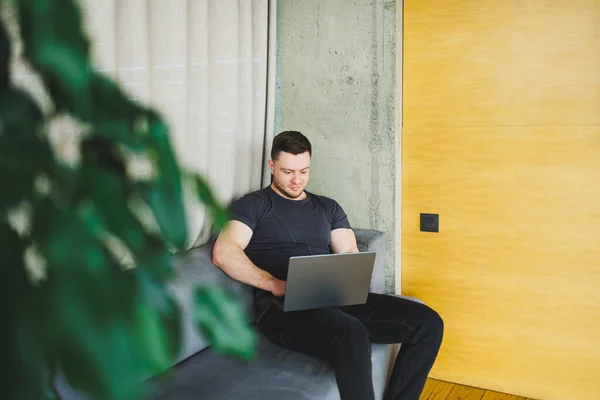 stock image Smiling man in t-shirt sitting on sofa, typing on netbook, working remotely on startup as freelancer, looking at laptop and smiling