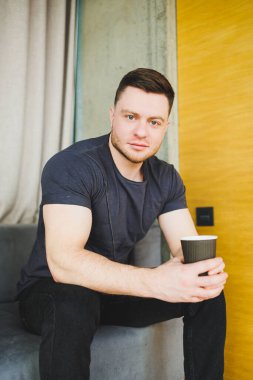 Tişörtlü yakışıklı genç adam kanepede oturuyor ve kahve içiyor, başlangıçta bağımsız olarak çalışıyor, dizüstü bilgisayarına bakıyor ve gülümsüyor.