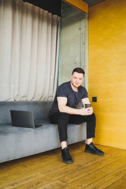 Tişörtlü yakışıklı genç adam kanepede oturuyor ve kahve içiyor, başlangıçta bağımsız olarak çalışıyor, dizüstü bilgisayarına bakıyor ve gülümsüyor.
