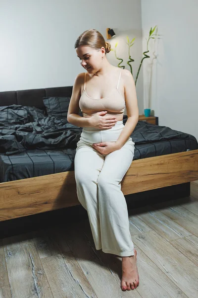 Cute young pregnant woman sitting on bed with gray sheets in casual clothes. Cute pregnant woman at home.