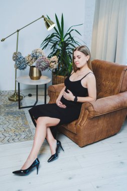 Attractive young blonde woman in black top and skirt sitting in brown chair and posing. Elegant woman in a soft chair.