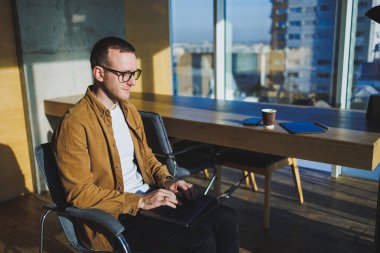 Geniş, modern bir ofiste uzaktan çalışan gülümseyen genç serbest çalışan bir erkek. Rahat giyimli bir yönetici ofiste rahat çalışır..