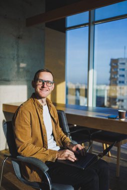 Geniş, modern bir ofiste uzaktan çalışan gülümseyen genç serbest çalışan bir erkek. Rahat giyimli bir yönetici ofiste rahat çalışır..