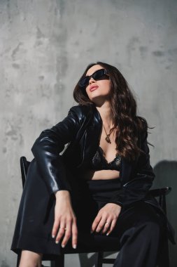 Deri ceketli, siyah pantolonlu, güneş gözlüklü genç bir kadın gri arka planda bir sandalyede oturuyor..
