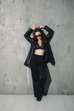 Gri arka planda poz veren zarif deri takım elbiseli, pantolon, ceket, şık güneş gözlüğü takan zarif genç kadın modeller..