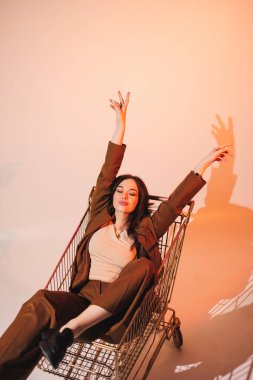 Gri arka planda alışveriş arabasında poz veren şık kahverengi takım elbiseli güzel, zarif genç bayan modeller. Takım elbiseli zarif bir bayan.