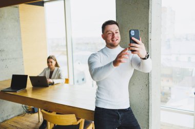 Beyaz kazaklı genç bir adam geniş bir ofiste duruyor ve telefonla konuşuyor. Bir adam video bağlantısı üzerinden iş arkadaşlarıyla konuşuyor.