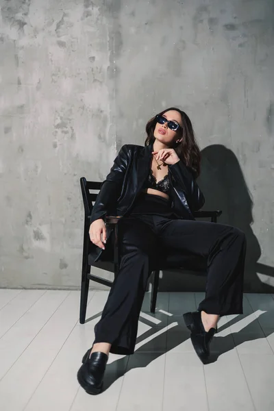A cute young woman in a leather jacket and black pants wearing sunglasses is sitting on a chair on a gray background.