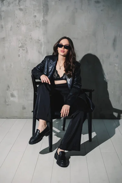 stock image A cute young woman in a leather jacket and black pants wearing sunglasses is sitting on a chair on a gray background.