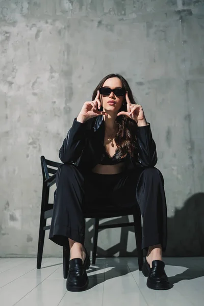 A cute young woman in a leather jacket and black pants wearing sunglasses is sitting on a chair on a gray background.