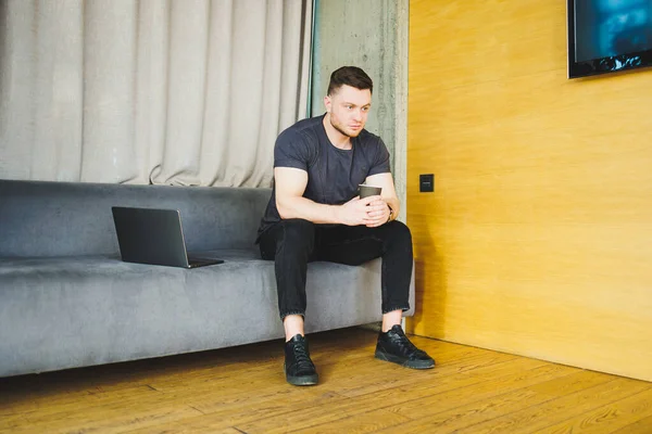 stock image Handsome young man in t-shirt sitting on sofa and drinking coffee, working remotely during startup as a freelancer, looking at laptop and smiling