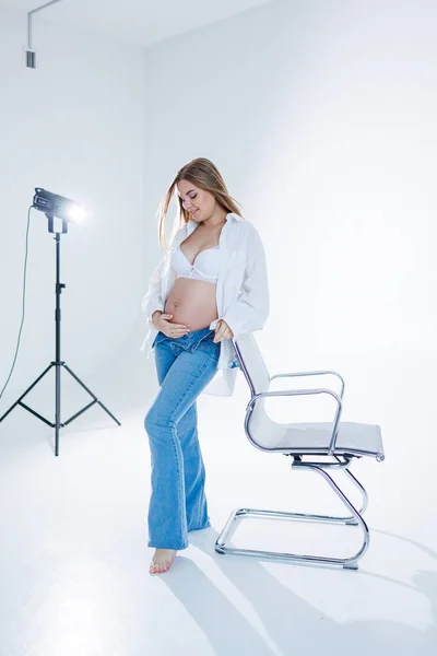 stock image Cute pregnant woman in bra and white shirt on white background. Happy pregnant woman stroking her belly.