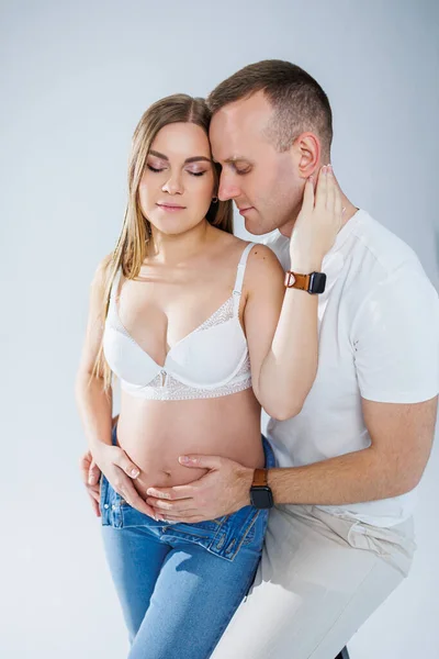 白い背景に白いTシャツとジーンズを着た赤ちゃんを期待して幸せな若いカップル 妊娠出産の幸せ 幸せな夫婦は子供を期待している — ストック写真