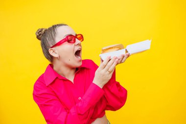 Pembe gömlekli ve kotlu hamile bir kadın lezzetli bir burger tutuyor. Hamilelik boyunca zararlı yiyecekler. Kolay ve mutlu hamilelik kavramı.