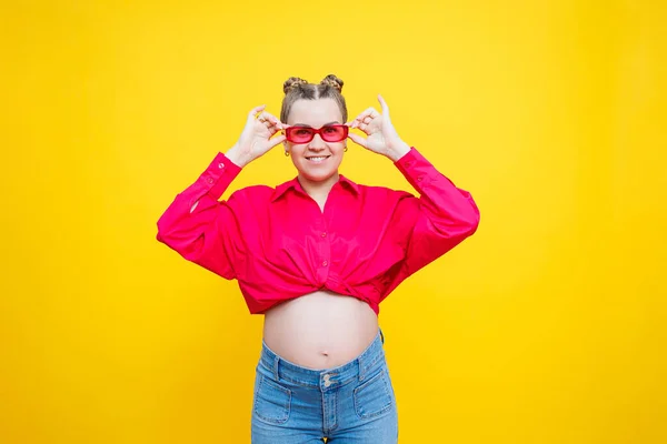 Happy pregnancy. Cheerful pretty pregnant woman in a pink shirt and pink glasses on a yellow background. Young bright pregnant woman.