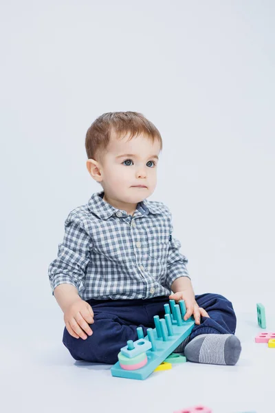 stock image A cute little boy in a checkered shirt sits on a white background and plays with wooden educational toys. Ecological wooden toys for children