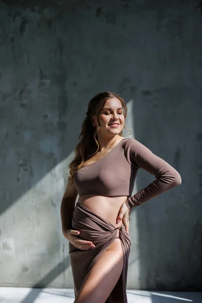 茶色の繊細なスーツを着た幸せな若い妊婦がポーズをとってお腹を撫でています 妊娠中の女性 幸せな妊娠 — ストック写真