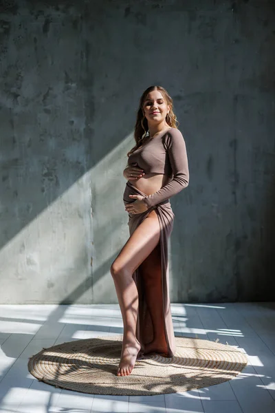茶色の繊細なスーツを着た幸せな若い妊婦がポーズをとってお腹を撫でています 妊娠中の女性 幸せな妊娠 — ストック写真