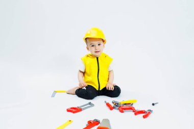 2 yaşında beyaz bir arka planda müteahhit kıyafeti giymiş küçük bir çocuk. Plastik kasklı ve plastik aletli bir çocuk. Plastik çocuk oyuncakları.