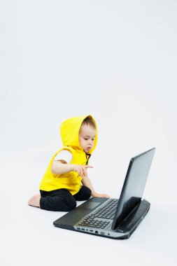 Sarı yelekli şirin bir çocuk beyaz arka planda dijital bir dizüstü bilgisayarın başında oturuyor. Çocuk ve modern taşınabilir bilgisayar aygıtı.