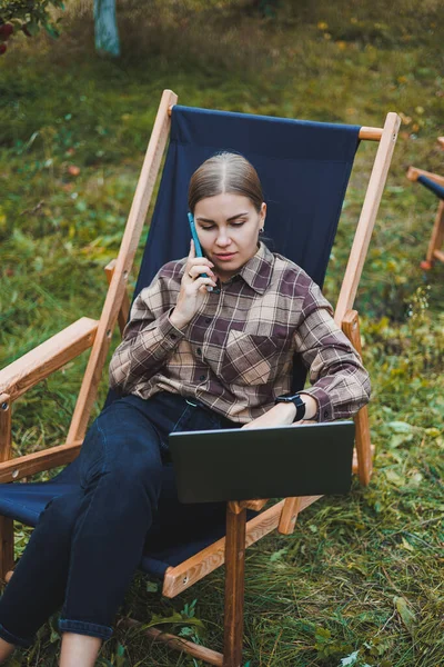 Stylish woman sitting on a chair near green plants in the garden and browsing the internet on a laptop while looking to the side. Remote work, female freelancer