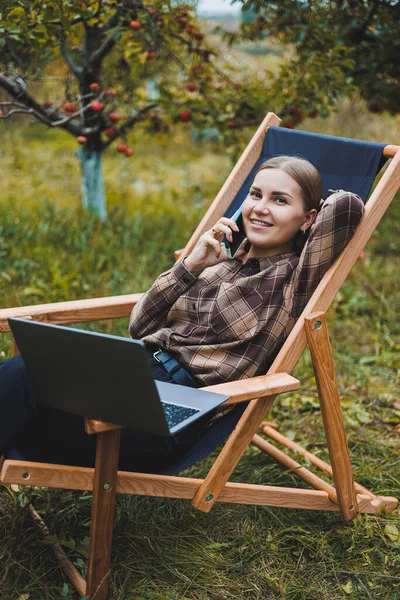 stock image Happy female freelancer working on digital netbook relaxing in garden chair and smiling at camera, woman with laptop posing in park during remote work