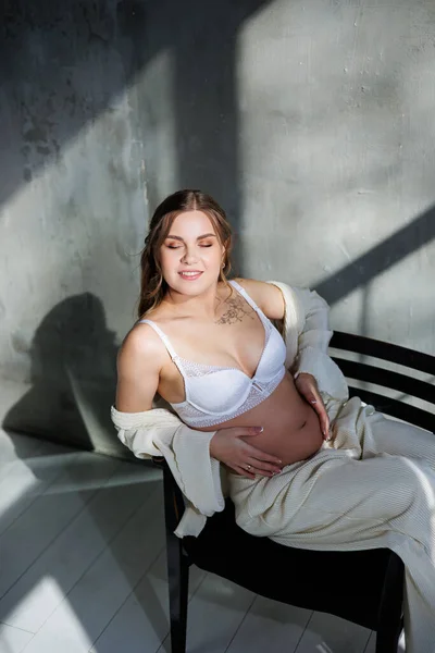 白いスーツとブラジャーを着た妊婦が灰色の背景に座っている 妊娠中の可愛い女性が腹を撫でて微笑んだ 妊娠の幸せな時期 — ストック写真