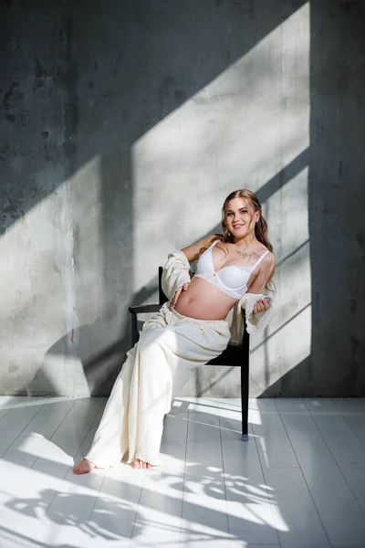 白髪の美しい女性が 灰色の背景の椅子に座っている白いエレガントなスーツに妊娠しています 妊婦のためのファッション スタイリッシュな女性は妊娠 — ストック写真