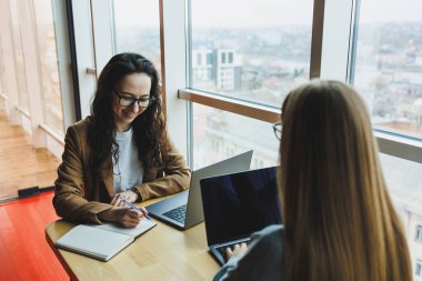 İki genç bayan meslektaşı büyük pencerelerin yanında dizüstü bilgisayarlarıyla oturuyorlar, modern çalışma alanı. Kadın ofis çalışanları