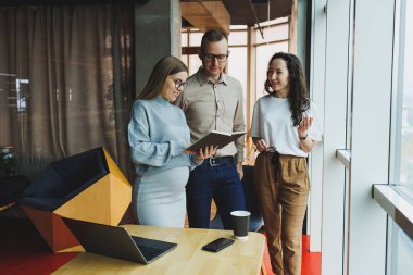 Ofiste iş arkadaşlarıyla çalışan modern hamile kadın. Genç ve başarılı bir adam hamile bir meslektaşına işte yardım ediyor. Güzel hamile bir kadın ofiste çalışıyor. Modern hamile kadın