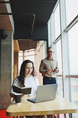 Gözlüklü bir kadın masada oturuyor ve dizüstü bilgisayarda çalışıyor. Arka planda çalışan bir erkek arkadaş telefonda konuşuyor. Modern bir ofiste çalışma günü..