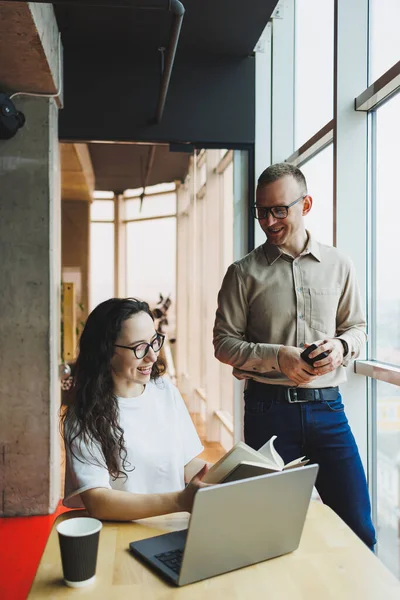 stock image A woman in glasses sits at a table and works on a laptop, a male colleague in the background is talking on the phone. A working day in a modern office.